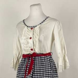 Puede incluir: Vestido blanco con falda de cuadros y corpiño de encaje. El corpiño tiene botones rojos y una cinta roja atada a la cintura.