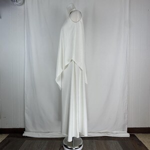 May include: A floor-length, white dress with a cape overlay. The dress is sleeveless with a high neckline and a flowing, elegant design. The cape drapes over the shoulders and down the back, creating a sophisticated silhouette. The dress is displayed on a mannequin.