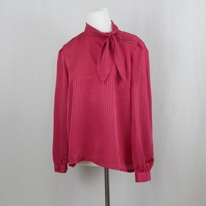 May include: A pink blouse with a gingham pattern and a bow tie at the neck. The blouse has long sleeves and a button closure at the shoulder.