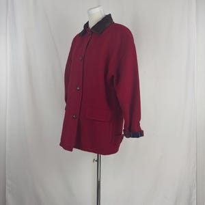 Op de afbeelding: Een bordeauxrode wollen jas met een bruine kraag en knoopsluiting. De jas heeft twee voorzakken en de manchetten zijn gevoerd met een blauw-zwart geruit patroon. De jas wordt op een mannequin getoond.