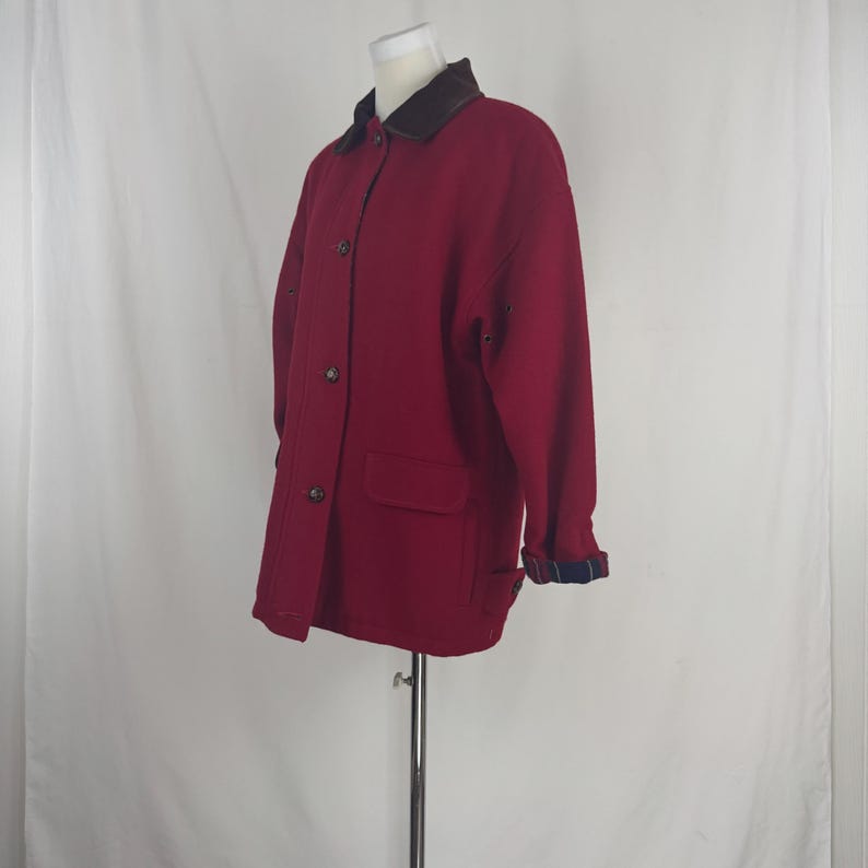 Op de afbeelding: Een bordeauxrode wollen jas met een bruine kraag en knoopsluiting. De jas heeft twee voorzakken en de manchetten zijn gevoerd met een blauw-zwart geruit patroon. De jas wordt op een mannequin getoond.