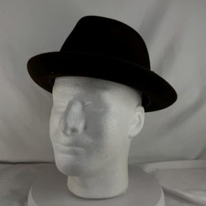 May include: A dark brown fedora hat with a slightly curved brim. The hat is displayed on a white mannequin head. The hat's crown is indented, and the brim is a consistent width around the hat.