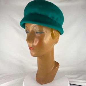May include: A green felt cloche hat with a rounded crown and a wide brim. The hat is sitting on a white mannequin head.