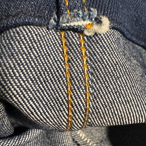 May include: Close-up of denim fabric with visible stitching. The image shows the inside and outside of the denim, with a focus on the seams and the contrasting orange thread used for the stitching. The fabric has a classic blue color.