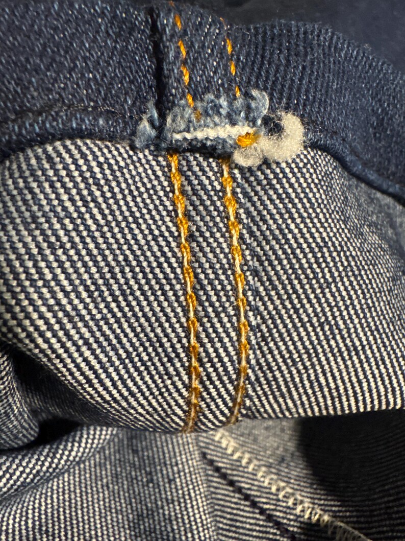 May include: Close-up of denim fabric with visible stitching. The image shows the inside and outside of the denim, with a focus on the seams and the contrasting orange thread used for the stitching. The fabric has a classic blue color.