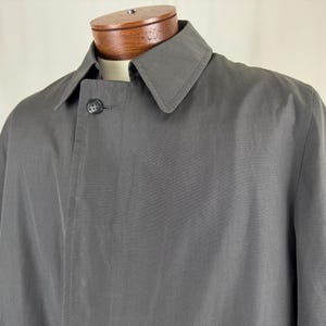 May include: A charcoal gray overcoat with a classic collar and a single button closure. The coat is displayed on a mannequin with a wooden neck and shoulders. The fabric appears to be a smooth, solid color.