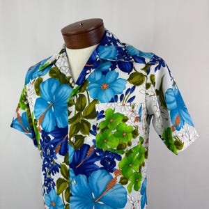 May include: A short-sleeved shirt with a vibrant floral pattern. The shirt features large blue hibiscus flowers, green leaves, and smaller white flowers against a white background. The shirt has a pocket on the chest and is buttoned up.