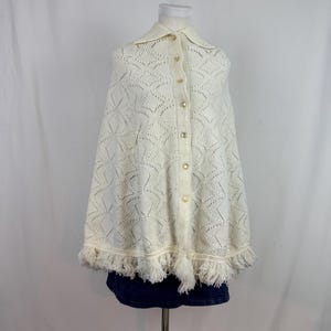 Op de afbeelding: Crèmekleurige gebreide cape met een kraag en knoopsluiting aan de voorkant. De cape heeft een geometrisch openwerkontwerp en een gefranjerde zoom. De knopen zijn hartvormig. Het kledingstuk wordt op een mannequin getoond.