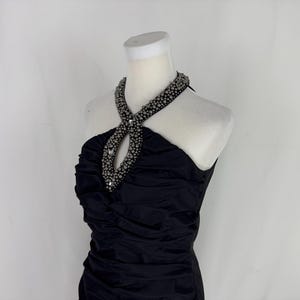 May include: A black, sleeveless dress with a ruched bodice and a halter neckline. The halter is embellished with silver-colored beads. The dress is displayed on a white mannequin against a white backdrop.