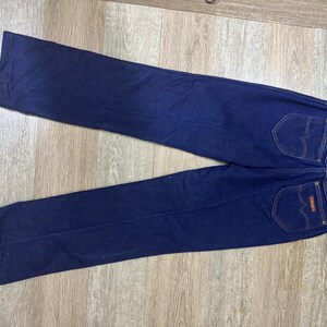 May include: Dark blue denim jeans with two back pockets and a small red and white label. The jeans are laid flat on a light brown wooden surface. The jeans have a classic, straight-leg design.