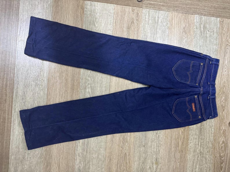May include: Dark blue denim jeans with two back pockets and a small red and white label. The jeans are laid flat on a light brown wooden surface. The jeans have a classic, straight-leg design.