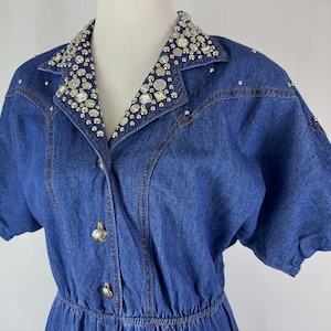 May include: A blue denim dress with a collared neckline and short sleeves. The collar is embellished with sparkling rhinestones. The dress features a button-down front and a cinched waist, creating a classic silhouette.