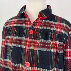May include: A close-up of a plaid jacket in red, navy, green, and white. The jacket has a collar, red buttons, and a red lining. The fabric appears to be a soft, woven material.