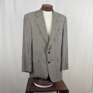 Könnte beinhalten: Ein Blazer mit Hahnentrittmuster in Schwarz und Beige, auf einer Schaufensterpuppe präsentiert. Die Jacke hat einen klassischen Zwei-Knopf-Verschluss, gekerbte Revers und Vordertaschen. Der Stoff hat eine subtile Textur und der Gesamtstil ist traditionell.