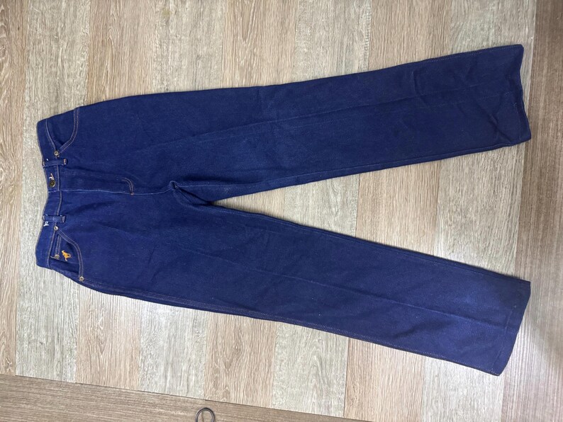 May include: Dark blue denim pants with a classic five-pocket design. The pants have a straight leg cut and a small embroidered horse logo on the front pocket. The pants are laid flat on a wooden surface.