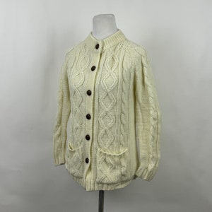 May include: A white cable knit cardigan with brown buttons and two patch pockets. The cardigan is on a white background.