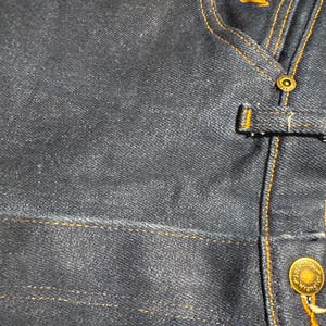 May include: Close-up of a dark blue denim jacket. The jacket features gold stitching, a button closure, and a small embroidered logo. The Wrangler brand name is visible on the button.