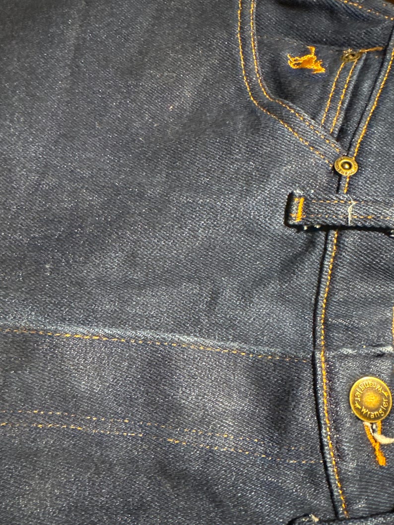 May include: Close-up of a dark blue denim jacket. The jacket features gold stitching, a button closure, and a small embroidered logo. The Wrangler brand name is visible on the button.
