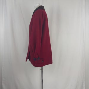 Op de afbeelding: Een bordeauxrode jas met een donkerbruine kraag en knoopsluitingen. De jas heeft lange mouwen en zakken. De manchetten hebben een blauw, groen en zwart ruitpatroon. De jas wordt op een mannequin gepresenteerd.