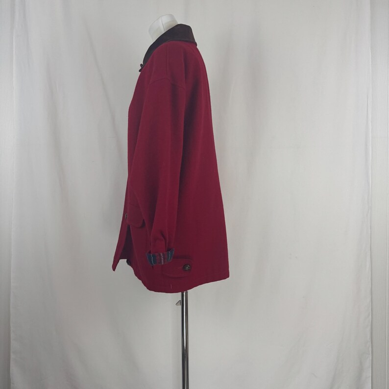 Op de afbeelding: Een bordeauxrode jas met een donkerbruine kraag en knoopsluitingen. De jas heeft lange mouwen en zakken. De manchetten hebben een blauw, groen en zwart ruitpatroon. De jas wordt op een mannequin gepresenteerd.