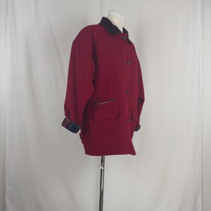Op de afbeelding: Een bordeauxrode jas met een zwarte kraag en knoopsluiting. De jas heeft twee voorzakken met ritsen en geruite manchetten. De jas wordt op een mannequin gepresenteerd.