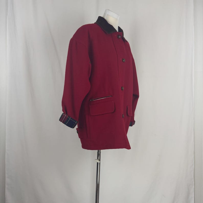 Op de afbeelding: Een bordeauxrode jas met een zwarte kraag en knoopsluiting. De jas heeft twee voorzakken met ritsen en geruite manchetten. De jas wordt op een mannequin gepresenteerd.