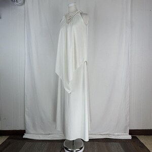 May include: A white, floor-length dress with a halter neckline and a flowing cape-like overlay. The neckline features a delicate, crocheted detail. The dress is displayed on a mannequin against a white backdrop, showcasing its elegant design.