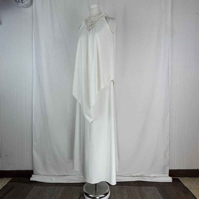 May include: A white, floor-length dress with a halter neckline and a flowing cape-like overlay. The neckline features a delicate, crocheted detail. The dress is displayed on a mannequin against a white backdrop, showcasing its elegant design.