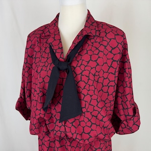 Op de afbeelding: Rode jurk met zwart patroon, gepresenteerd op een mannequin. De jurk heeft een kraag, lange mouwen en een zwarte stropdas. De stof heeft een herhalend patroon van zwarte vormen op een rode achtergrond.