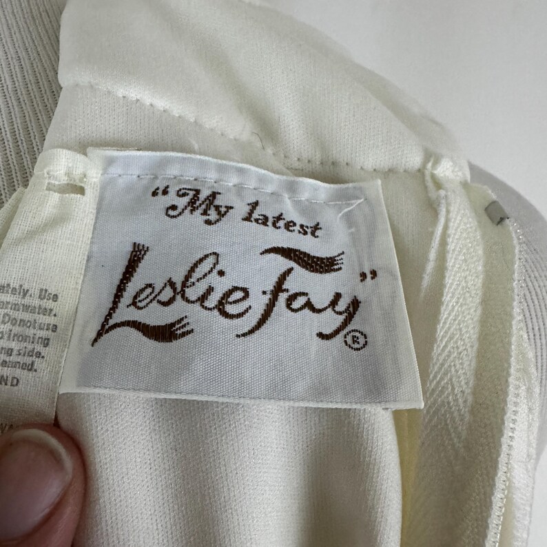 May include: Close-up of a cream-colored garment with a white label. The label reads "My latest Leslie Fay" in brown script. The garment has a zipper and appears to be made of a textured fabric. The overall aesthetic is vintage.