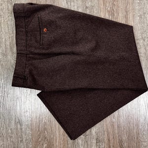 May include: Dark brown trousers with a buttoned pocket detail. The pants are made of a textured fabric and are folded neatly. The garment is displayed on a wooden surface.