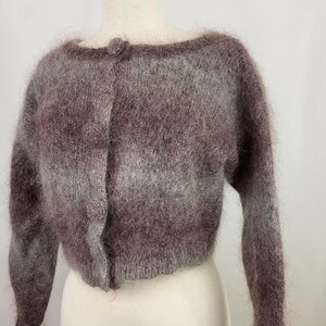 May include: A cropped, long-sleeved cardigan made of a fuzzy, brushed knit fabric in a variegated blend of muted brown, mauve, and grey tones. The garment features a round neckline, a front button closure with matching buttons, and ribbed cuffs.