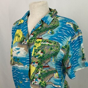 May include: A short-sleeved button-down shirt featuring a vibrant blue tropical coastal print. The pattern displays yellow cliffs, green palm trees, white buildings with red roofs, and small boats on the water. It has a classic collar and buttons.