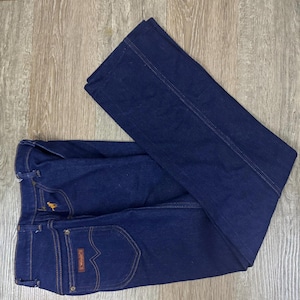 May include: Dark blue denim jeans with a classic design. Features include a button and zipper closure, front and back pockets, and a small embroidered logo. The jeans are folded on a wooden surface.