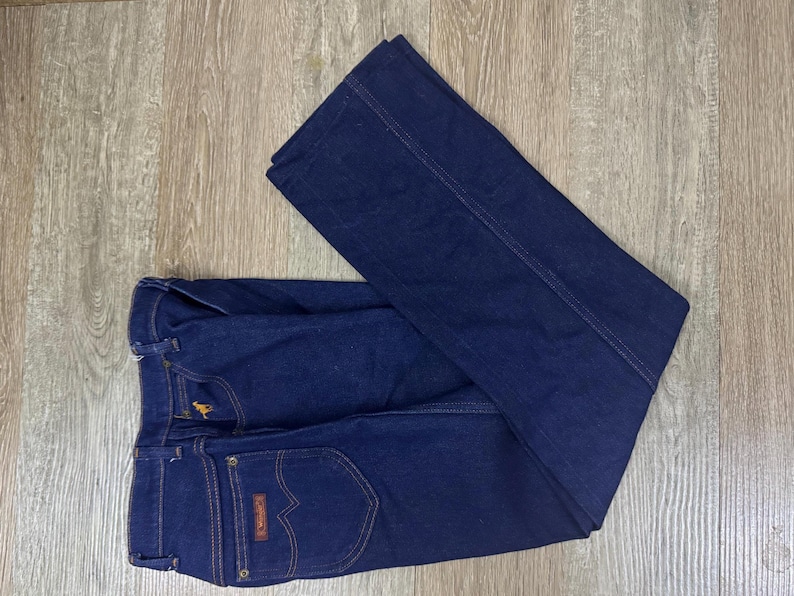 May include: Dark blue denim jeans with a classic design. Features include a button and zipper closure, front and back pockets, and a small embroidered logo. The jeans are folded on a wooden surface.