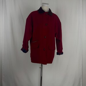 Op de afbeelding: Een rode jas met een donkerbruine kraag en geruite manchetten. De jas heeft twee voorzakken en een knoopsluiting. De mouwen zijn lang en de jas lijkt van een dik materiaal te zijn.