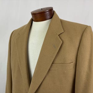 May include: A tan blazer displayed on a mannequin. The blazer has a notched lapel, a single-breasted closure, and a chest pocket. The fabric has a subtle texture. The blazer is a neutral color, suitable for various outfits.