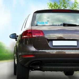 May include: A brown SUV with the text "Psalm 91" in light blue on the rear window. The vehicle is on a road with a blurred background of sky and trees. The rear lights are red.