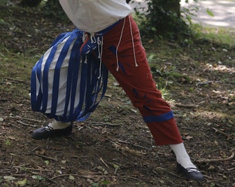 Op de afbeelding: Een man in een wit shirt en een rode en blauwe broek houdt een zwaard vast en poseert in een bos.