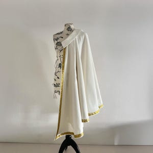 May include: A white garment with gold trim is draped over a mannequin. The mannequin's torso is covered in a black and white floral pattern. The garment is a flowing, elegant design.