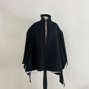 May include: A black cape-style jacket with a high collar and a front zipper. The jacket has a wide, flowing silhouette and is displayed on a black mannequin. The interior lining is a light color with a subtle pattern.