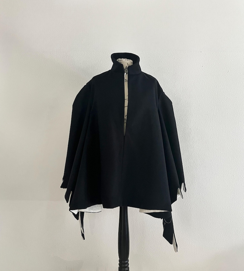 May include: A black cape-style jacket with a high collar and a front zipper. The jacket has a wide, flowing silhouette and is displayed on a black mannequin. The interior lining is a light color with a subtle pattern.
