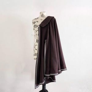 May include: A dark brown cape with white trim, draped over a floral patterned dress form. The cape has a wide collar and a flowing design, with white stripes along the edges.