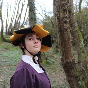 Puede incluir: Una persona vestida con un vestido morado y un sombrero negro y amarillo de ala ancha. El sombrero tiene un dise&ntilde;o &uacute;nico con m&uacute;ltiples puntos. La persona est&aacute; de pie en un &aacute;rea boscosa.