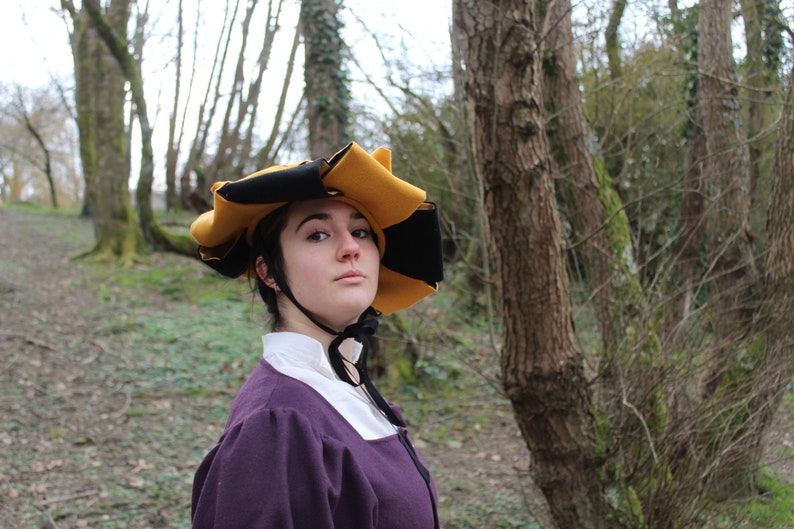 Puede incluir: Una persona vestida con un vestido morado y un sombrero negro y amarillo de ala ancha. El sombrero tiene un dise&ntilde;o &uacute;nico con m&uacute;ltiples puntos. La persona est&aacute; de pie en un &aacute;rea boscosa.