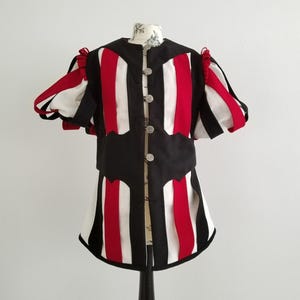 Pode incluir: Um gibão de estilo medieval com um design listrado em preto, vermelho e branco. A peça de manga curta apresenta botões prateados decorativos e fitas vermelhas nos ombros. A parte inferior do gibão tem uma borda ondulada.