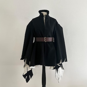 May include: A black cape-style coat with a high collar and a brown belt. The coat features white, leaf-shaped accents at the sleeves and hem. The coat is displayed on a black mannequin.