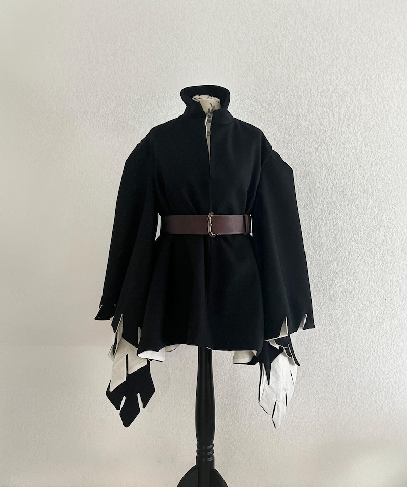 May include: A black cape-style coat with a high collar and a brown belt. The coat features white, leaf-shaped accents at the sleeves and hem. The coat is displayed on a black mannequin.
