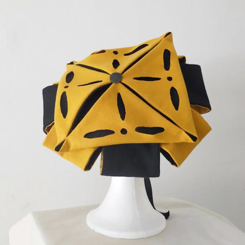 Puede incluir: Un sombrero amarillo con recortes negros y una cinta negra. El sombrero est&aacute; en un soporte de exhibici&oacute;n blanco.