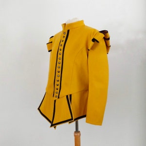Puede incluir: Una chaqueta de color amarillo mostaza con ribetes negros y botones decorativos. La chaqueta tiene un cuello alto y mangas largas. La parte inferior de la chaqueta está cortada en forma de punta.